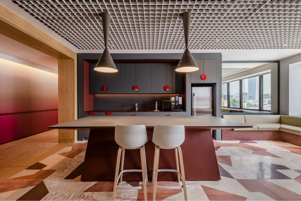 A modern office kitchen pantry with a central island, two white bar stools, and distinctive designer lighting.